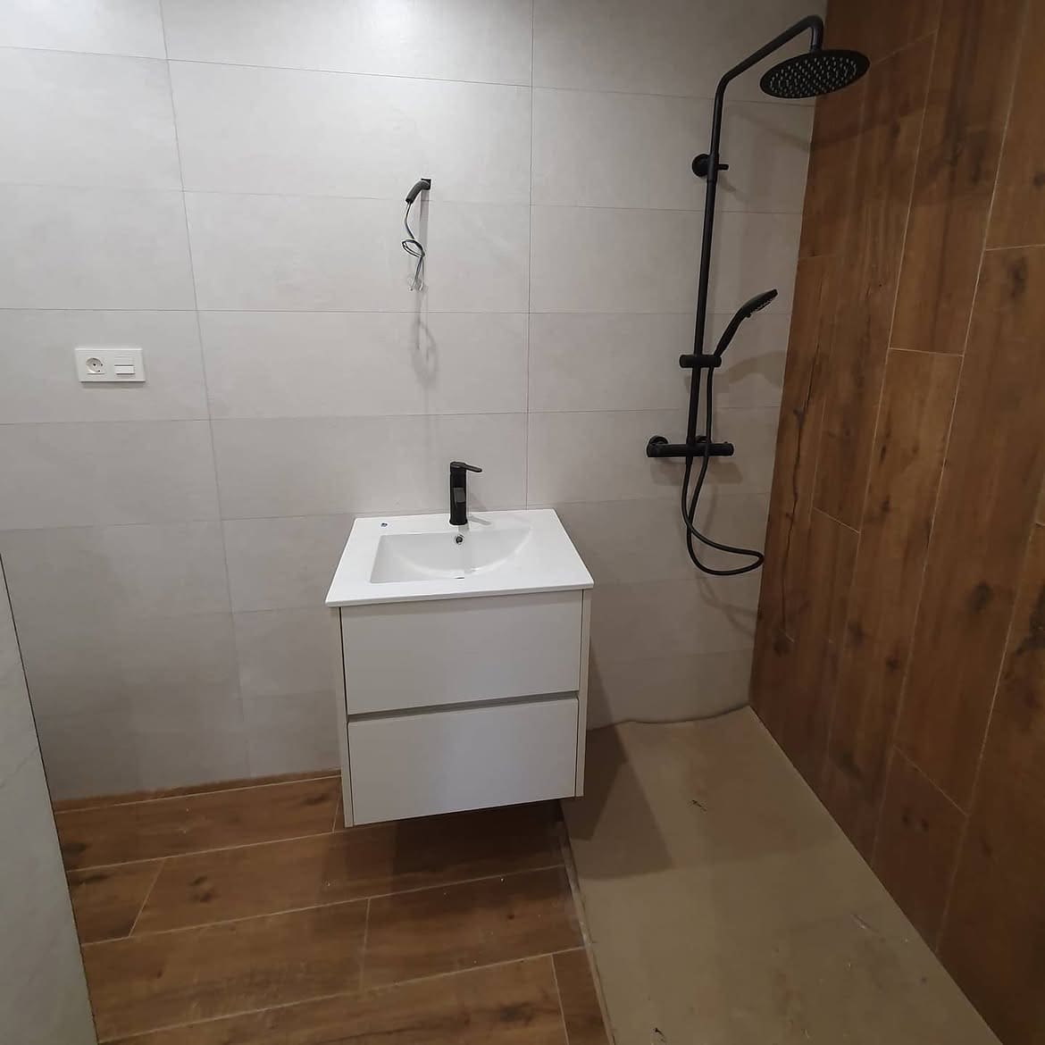 Baño en fase de acabados con lavabo suspendido blanco, grifería y columna de ducha en negro mate y revestimiento de gres gris combinado con gres efecto madera.