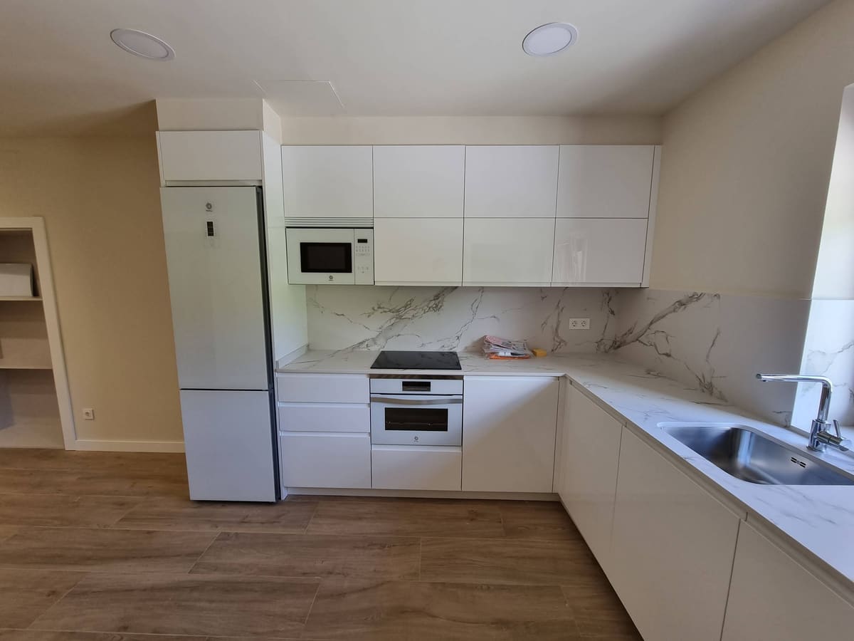 Cocina en L con muebles blancos sin tiradores, encimera y frontal de porcelánico efecto mármol, placa de inducción, horno y microondas integrados y suelo de tono madera.