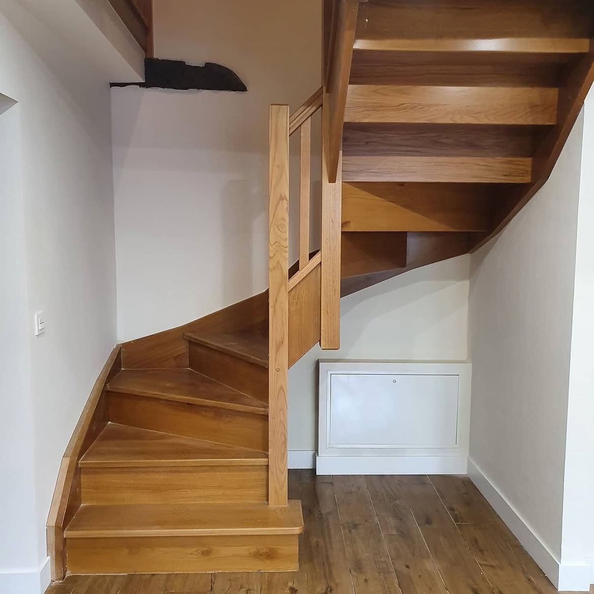 Escalera de madera con peldaños compensados y barandilla de madera, junto a un registro de instalaciones bajo la escalera.