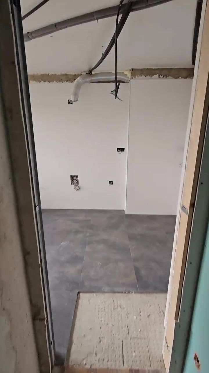 Fotograma del vídeo: cocina en obra con instalaciones vistas en el techo, tomas en la pared y pavimento cerámico gris ya colocado.