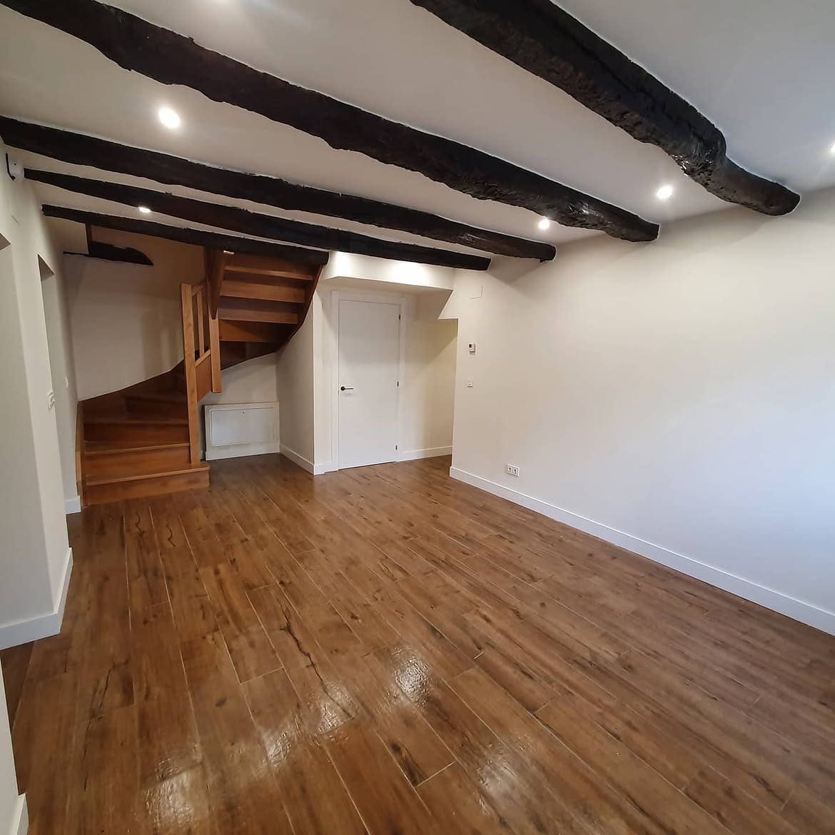 Salón reformado con vigas de madera oscura vistas, focos empotrados, paredes blancas, suelo de tono madera y escalera de madera al fondo.